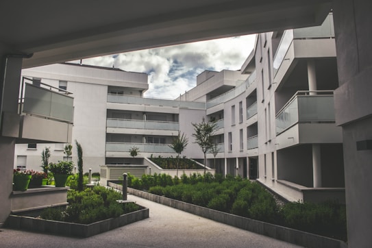 A modern residential complex featuring curved architecture with multiple stories. The buildings are white, with glass balustrades on the balconies. The setting includes landscaped green areas with well-maintained shrubs and small trees. The sky is partially cloudy, creating a serene and tranquil environment.