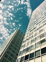 Two tall modern skyscrapers with numerous windows reach up into the sky, surrounded by fluffy white clouds against a blue backdrop.