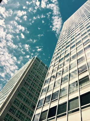 Two tall modern skyscrapers with numerous windows reach up into the sky, surrounded by fluffy white clouds against a blue backdrop.