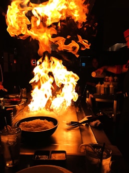 A dramatic display of fire rises from a teppanyaki grill, illuminating the dimly lit restaurant setting. A chef, partially visible to the right, appears to be skillfully handling the flames using a bottle. Plates and drinks are arranged on the table, and a bowl of rice is prominently placed near the grill.