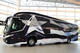 A sleek, modern bus is parked indoors with a unique black and white pattern on its exterior. The bus displays logos and appears to be customized, likely for promotional or team transportation purposes. Large windows and aerodynamic design convey a sense of luxury.