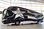 A sleek, modern bus is parked indoors with a unique black and white pattern on its exterior. The bus displays logos and appears to be customized, likely for promotional or team transportation purposes. Large windows and aerodynamic design convey a sense of luxury.