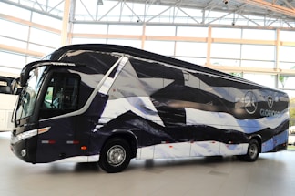 A sleek, modern bus is parked indoors with a unique black and white pattern on its exterior. The bus displays logos and appears to be customized, likely for promotional or team transportation purposes. Large windows and aerodynamic design convey a sense of luxury.