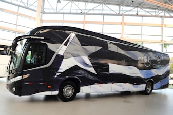A sleek, modern bus is parked indoors with a unique black and white pattern on its exterior. The bus displays logos and appears to be customized, likely for promotional or team transportation purposes. Large windows and aerodynamic design convey a sense of luxury.