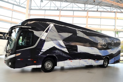A sleek, modern bus is parked indoors with a unique black and white pattern on its exterior. The bus displays logos and appears to be customized, likely for promotional or team transportation purposes. Large windows and aerodynamic design convey a sense of luxury.