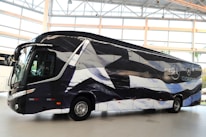 A sleek, modern bus is parked indoors with a unique black and white pattern on its exterior. The bus displays logos and appears to be customized, likely for promotional or team transportation purposes. Large windows and aerodynamic design convey a sense of luxury.