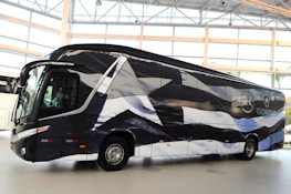 A sleek, modern bus is parked indoors with a unique black and white pattern on its exterior. The bus displays logos and appears to be customized, likely for promotional or team transportation purposes. Large windows and aerodynamic design convey a sense of luxury.