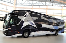 A sleek, modern bus is parked indoors with a unique black and white pattern on its exterior. The bus displays logos and appears to be customized, likely for promotional or team transportation purposes. Large windows and aerodynamic design convey a sense of luxury.