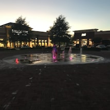 A plaza with a fountain featuring colorful lights illuminating the water streams, situated in front of a shopping center. The surrounding area includes trees and warmly lit storefronts, with a few parked cars visible. The atmosphere suggests evening as the sky is darkening.