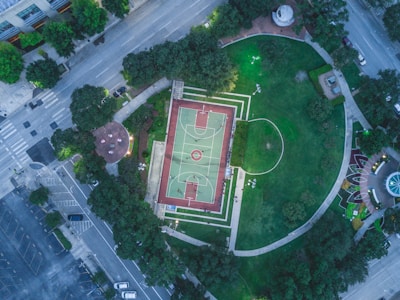 An aerial view of a vibrant urban park featuring a basketball court at the center surrounded by lush greenery. The park is bordered by streets and parking areas, with visible pathways, benches, and a small round seating area with tables. The well-maintained grass and decorative landscaping enhance the scene.