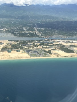 Aerial view of a coastal area featuring a mix of urban development and natural landscapes. The foreground shows a stretch of blue ocean meeting a sandy beach. Further inland, there is a residential or resort area with clusters of buildings surrounded by greenery. Roads and infrastructure are visible, connecting the developed areas. Beyond, the landscape transitions into lush green fields and mountainous terrain under a sky filled with scattered clouds.