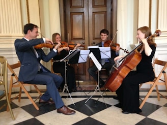 A quartet of musicians is playing string instruments in an elegant room. The group consists of two people playing violins, one on viola, and another on a cello. The musicians are seated on wooden chairs arranged in a semi-circle, and music stands with sheet music are placed in front of them. The flooring is checkered, and the room features classic wooden doors and ornate wall details.
