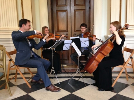 A quartet of musicians is playing string instruments in an elegant room. The group consists of two people playing violins, one on viola, and another on a cello. The musicians are seated on wooden chairs arranged in a semi-circle, and music stands with sheet music are placed in front of them. The flooring is checkered, and the room features classic wooden doors and ornate wall details.