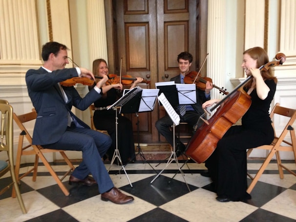 A quartet of musicians is playing string instruments in an elegant room. The group consists of two people playing violins, one on viola, and another on a cello. The musicians are seated on wooden chairs arranged in a semi-circle, and music stands with sheet music are placed in front of them. The flooring is checkered, and the room features classic wooden doors and ornate wall details.