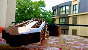 A close-up view of a guitar headstock resting on a table with a patterned cloth, set against the backdrop of an urban residential building and green trees.