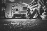 In a dimly lit garage, a person lies on the ground working underneath a vintage Volkswagen Beetle. The garage is cluttered with various tools, shelves packed with containers, and car-related items, creating a detailed and authentic mechanical environment.
