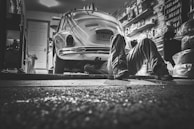 In a dimly lit garage, a person lies on the ground working underneath a vintage Volkswagen Beetle. The garage is cluttered with various tools, shelves packed with containers, and car-related items, creating a detailed and authentic mechanical environment.