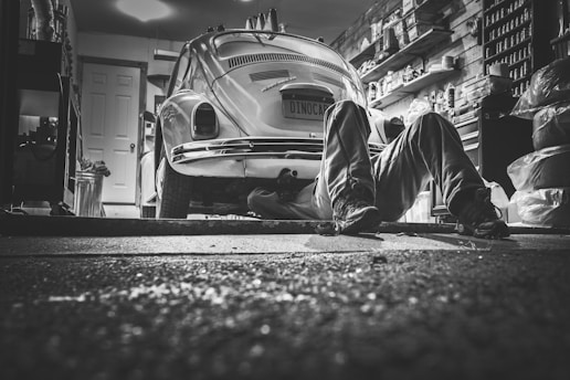 In a dimly lit garage, a person lies on the ground working underneath a vintage Volkswagen Beetle. The garage is cluttered with various tools, shelves packed with containers, and car-related items, creating a detailed and authentic mechanical environment.