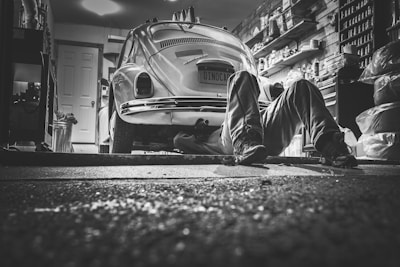 In a dimly lit garage, a person lies on the ground working underneath a vintage Volkswagen Beetle. The garage is cluttered with various tools, shelves packed with containers, and car-related items, creating a detailed and authentic mechanical environment.