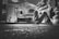 In a dimly lit garage, a person lies on the ground working underneath a vintage Volkswagen Beetle. The garage is cluttered with various tools, shelves packed with containers, and car-related items, creating a detailed and authentic mechanical environment.