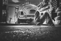 In a dimly lit garage, a person lies on the ground working underneath a vintage Volkswagen Beetle. The garage is cluttered with various tools, shelves packed with containers, and car-related items, creating a detailed and authentic mechanical environment.