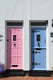 Two adjacent doors are set in a white wall, one painted pink and the other blue. Each door features ornate black metalwork, including door handles and decorative hinges. A small number '6' is displayed on the pink door, and there is a brick walkway at the base.