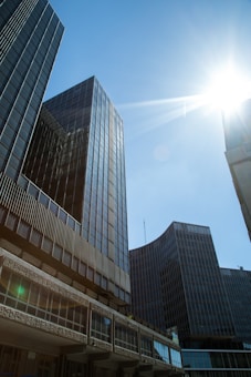 Tall modern office buildings with reflective glass facades rise against a clear blue sky. The sun shines brightly in the top right corner, casting lens flares and shadows on the structures.