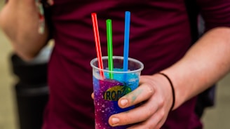 A person holding a cup filled with a vibrant blue and purple slushy drink. The drink is adorned with three colorful straws in red, green, and blue. The person is wearing a burgundy shirt, and there is a hint of a black bracelet on their wrist.