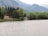 A serene lakeside scene with a small house featuring a yellow facade near the shore, surrounded by dense green trees. A quaint boathouse is partially hidden by the foliage, and the lake is calm with a swan gliding across the water. Mountains rise in the background under a cloudy sky.