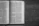 An open book lies on a wooden surface, displaying pages filled with dense text in a serif font. The book appears to be well-worn, with slightly curved pages and a textured cover. The wooden surface beneath has a rough, natural finish with visible grain patterns.