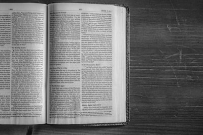 An open book lies on a wooden surface, displaying pages filled with dense text in a serif font. The book appears to be well-worn, with slightly curved pages and a textured cover. The wooden surface beneath has a rough, natural finish with visible grain patterns.