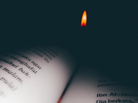 A small flame provides light above an open book with text in a dark setting. The words are partially obscured by shadows, creating a cozy and intimate atmosphere.