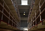 A long corridor flanked by multiple levels of barred prison cells, with red accents on the railings. The lighting creates stark shadows, and there is a clock visible in the distance on the wall.