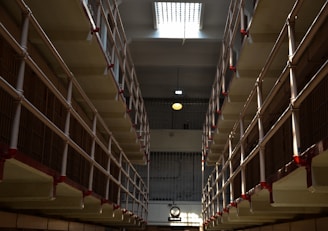 A long corridor flanked by multiple levels of barred prison cells, with red accents on the railings. The lighting creates stark shadows, and there is a clock visible in the distance on the wall.