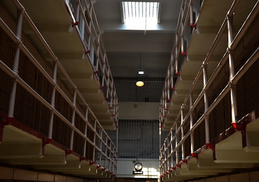 A long corridor flanked by multiple levels of barred prison cells, with red accents on the railings. The lighting creates stark shadows, and there is a clock visible in the distance on the wall.