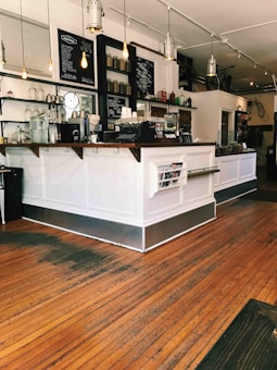 A cozy coffee shop interior featuring a white counter with wooden accents, a blackboard menu, and shelves stocked with glass bottles and jars. Warm lighting from hanging bulbs creates an inviting atmosphere, and the wooden floor adds to the rustic charm. In the background, a bicycle is mounted on the wall as part of the decor.