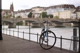 A bicycle is parked against a railing beside a river with a view of a historic cityscape in the background. The scene includes a stone bridge crossing the river, lined with buildings featuring classic European architectural styles, and a slightly overcast sky.