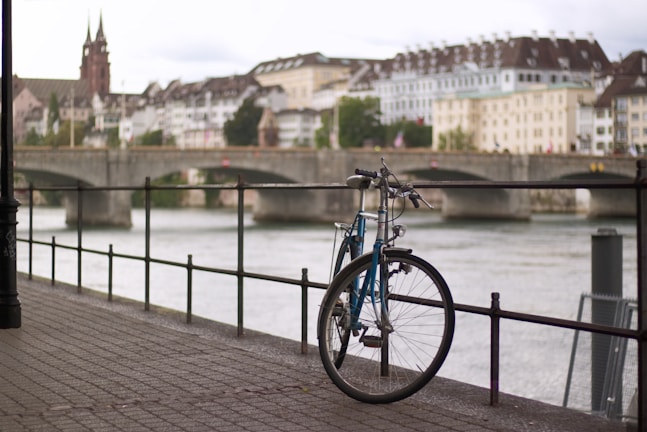 A bicycle is parked against a railing beside a river with a view of a historic cityscape in the background. The scene includes a stone bridge crossing the river, lined with buildings featuring classic European architectural styles, and a slightly overcast sky.