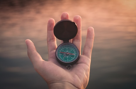 A hand holds a compass with a green dial against a soft-focus background of a sunset over water. The compass is open, and its needle is visible. The lighting is warm, casting a gentle glow on the hand and the compass.