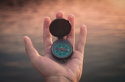 A hand holds a compass with a green dial against a soft-focus background of a sunset over water. The compass is open, and its needle is visible. The lighting is warm, casting a gentle glow on the hand and the compass.