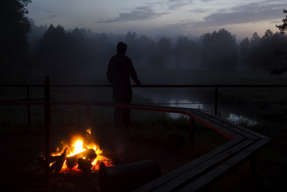 silhouette photo of a person standing near body of water and bonfire