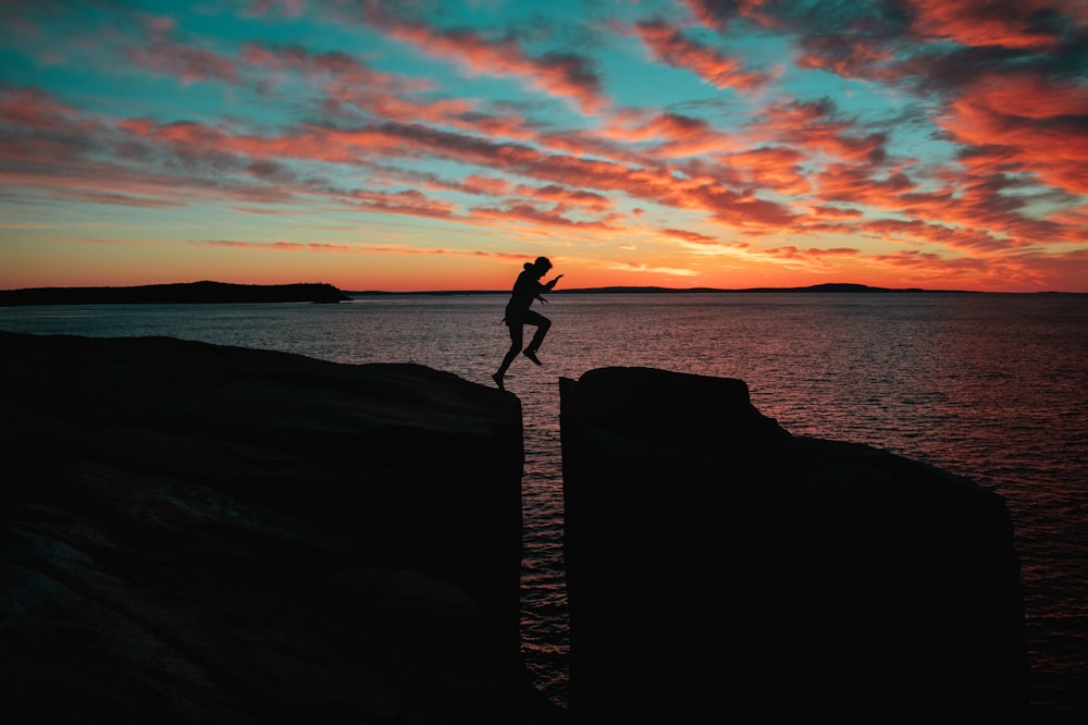 日没時に海の近くの崖に飛び乗る男のシルエット写真