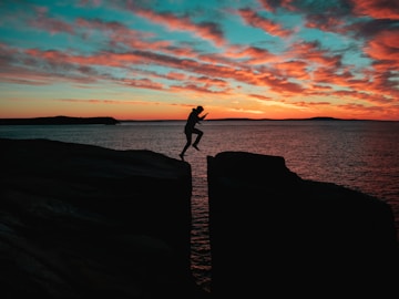 silhouette photo of a man jumps on to cliff near sea during sunset