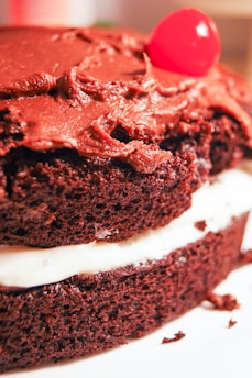 A close-up view of a chocolate layer cake with rich, creamy chocolate icing on the top. The cake is layered with a white filling, adding contrast to the dark chocolate sponge. A single bright red cherry sits atop the cake, providing a pop of color.