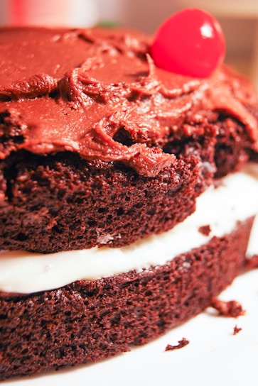 A close-up view of a chocolate layer cake with rich, creamy chocolate icing on the top. The cake is layered with a white filling, adding contrast to the dark chocolate sponge. A single bright red cherry sits atop the cake, providing a pop of color.