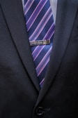 A close-up of a person wearing a dark suit jacket with a tie featuring diagonal stripes in blue, pink, and purple shades. A tie clip reading 'Grandpa' is attached to the tie.