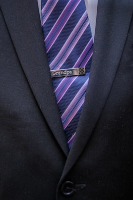A close-up of a person wearing a dark suit jacket with a tie featuring diagonal stripes in blue, pink, and purple shades. A tie clip reading 'Grandpa' is attached to the tie.