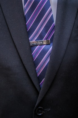 A close-up of a person wearing a dark suit jacket with a tie featuring diagonal stripes in blue, pink, and purple shades. A tie clip reading 'Grandpa' is attached to the tie.