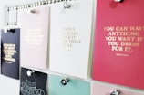 A collection of motivational quotes displayed on different colored cards pinned to a board. Each card has gold lettering featuring different inspirational sayings. The board is organized with silver pins holding the cards in place.