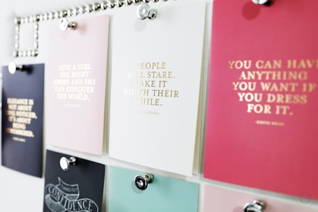 A collection of motivational quotes displayed on different colored cards pinned to a board. Each card has gold lettering featuring different inspirational sayings. The board is organized with silver pins holding the cards in place.